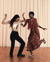 Two women dancing on a wooden floor in front of pale pleated curtains for the Rabanne Resort 2026 party collection.