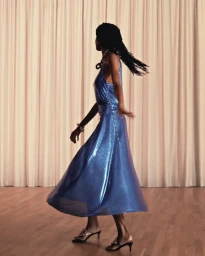 Model in motion wearing a shiny blue dress and heels against pleated curtains