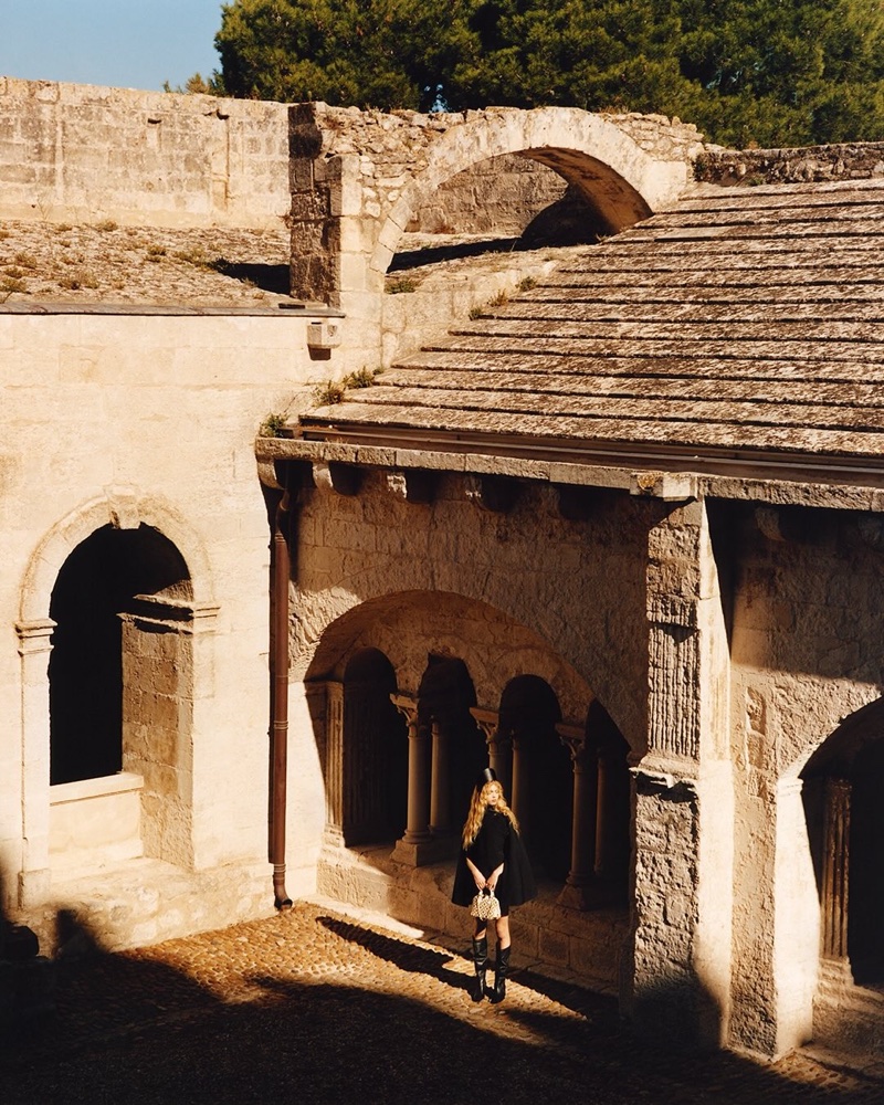 Model with blonde hair wearing a black dress and boots while holding a handbag in a sunlit stone courtyard.
