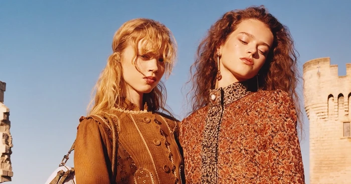 Two female models in ornate patterned garments outdoors against a clear blue sky with a stone architectural tower in the background.