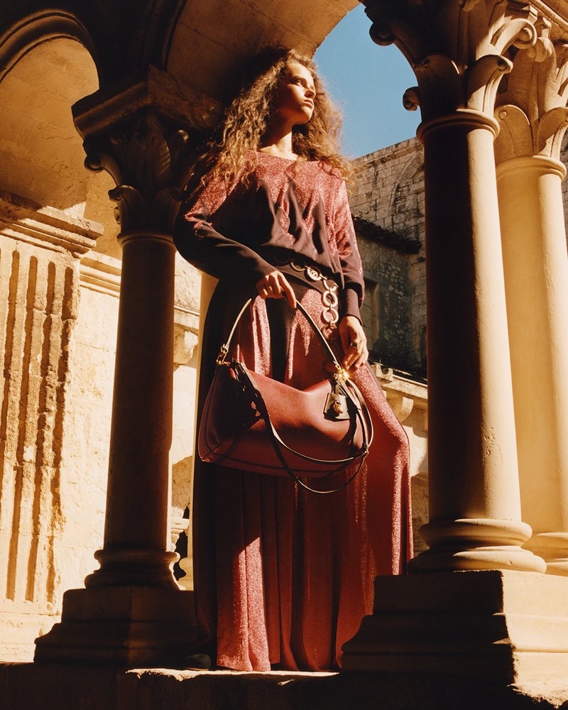 Model standing between stone architectural columns holding a leather shoulder bag in bright sunlight