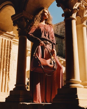 Model standing between stone architectural columns holding a leather shoulder bag in bright sunlight