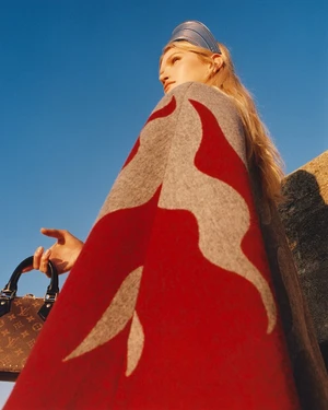 Model wearing a grey and red patterned cape and metallic visor holding a monogram handbag against a bright blue sky.