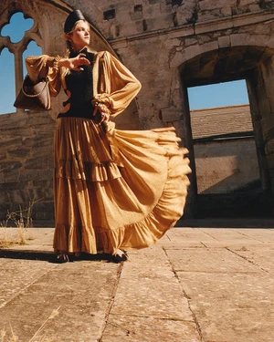 Model in a golden ruffled maxi skirt and black top holding a handbag before weathered stone architectural ruins.