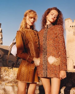 Two female models in patterned designer coats posing outdoors against a historic stone building under clear sky.