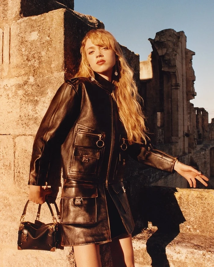 Model with long blonde hair wearing a black leather jacket and skirt, holding a handbag against stone ruins.