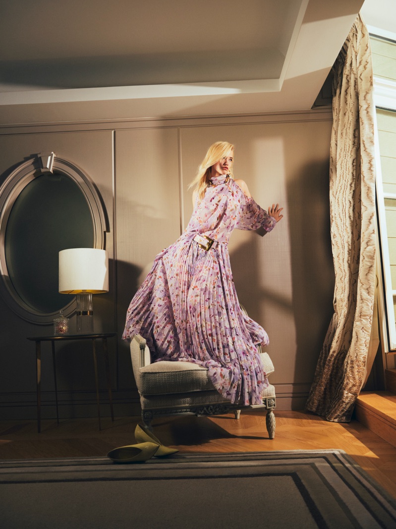 Blonde woman in a printed lilac pleated gown standing barefoot on an upholstered armchair beside an oval mirror and window drapes.