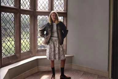 Gigi Hadid standing indoors in front of large leaded glass windows wearing a dark leather jacket and patterned skirt.