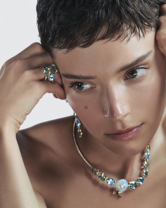 Close-up portrait of a short-haired model wearing gemstone rings and a diamond and colored stone necklace against a plain white background.