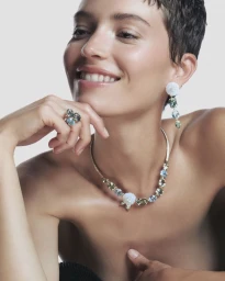 Model with short dark hair smiling while wearing gem-encrusted necklace, floral earrings, and ring against a plain white background.