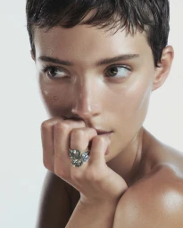Close-up portrait of a woman with short dark hair wearing a large statement ring against a white background