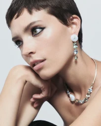 Close-up portrait of a woman with short dark hair wearing ornate gemstone earrings and a statement necklace against a light grey background.