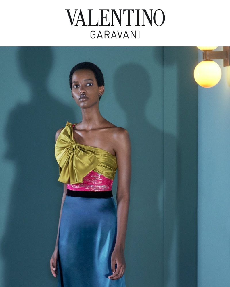 Model wearing a one-shoulder yellow bow bodice and blue satin skirt against a teal wall with wall sconce