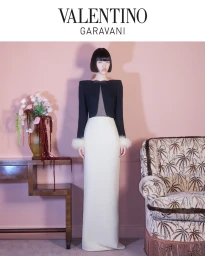 Model in a black and white gown with feathered cuffs standing in a pink room with a patterned armchair.