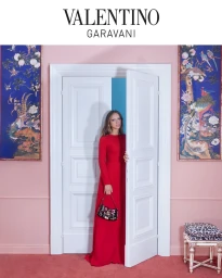 Model in a long red dress holding a patterned handbag standing in a doorway between pink walls and blue framed artwork