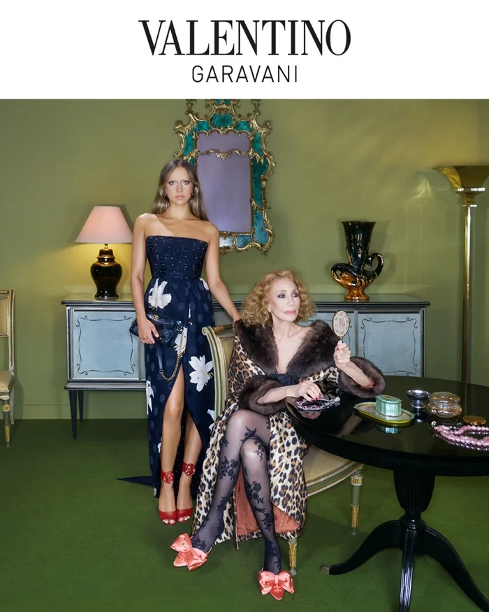 Two women in an olive green room, one standing in a blue strapless gown and one seated wearing an animal print coat.
