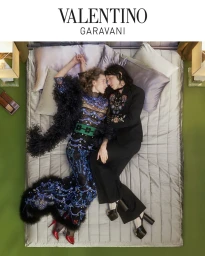 Two people lying on a quilted grey bed surrounded by green flooring, wearing embroidered Valentino fashion apparel.