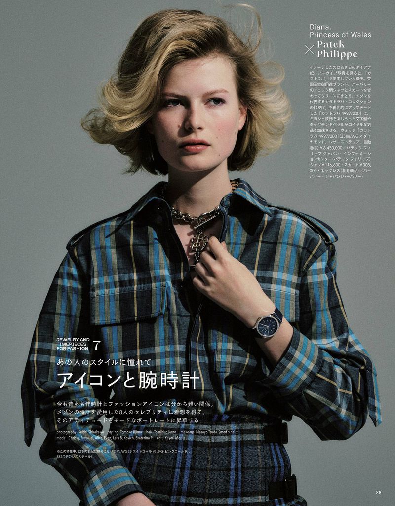 A young woman with blonde hair poses indoors wearing a blue plaid shirt, statement necklace, and wristwatch against a grey backdrop.