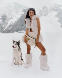 Emily Ratajkowski in white faux fur vest and boots holding a furry handbag with a husky on snow for Kurt Geiger Holiday 2025 by Laura Coulson