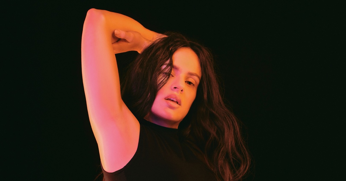 Rosalia posing in a black sleeveless top against a dark background with warm pink and orange studio lighting.