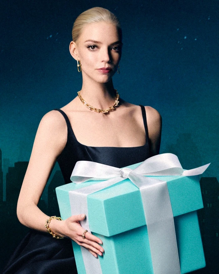 Anya Taylor-Joy wearing a gold necklace and holding a large Tiffany blue gift box with a white ribbon against a night cityscape background.