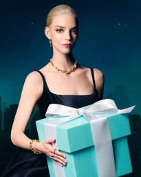 Anya Taylor-Joy wearing a gold necklace and holding a large Tiffany blue gift box with a white ribbon against a night cityscape background.