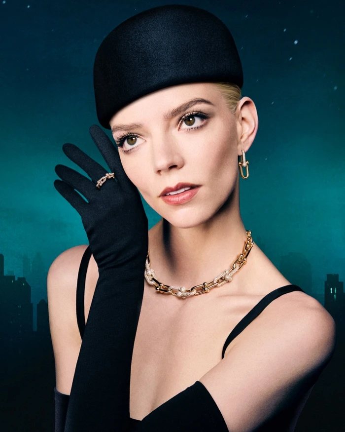 Anya Taylor-Joy wearing a black pillbox hat, black gloves, and gold jewellery against a dark teal cityscape background.