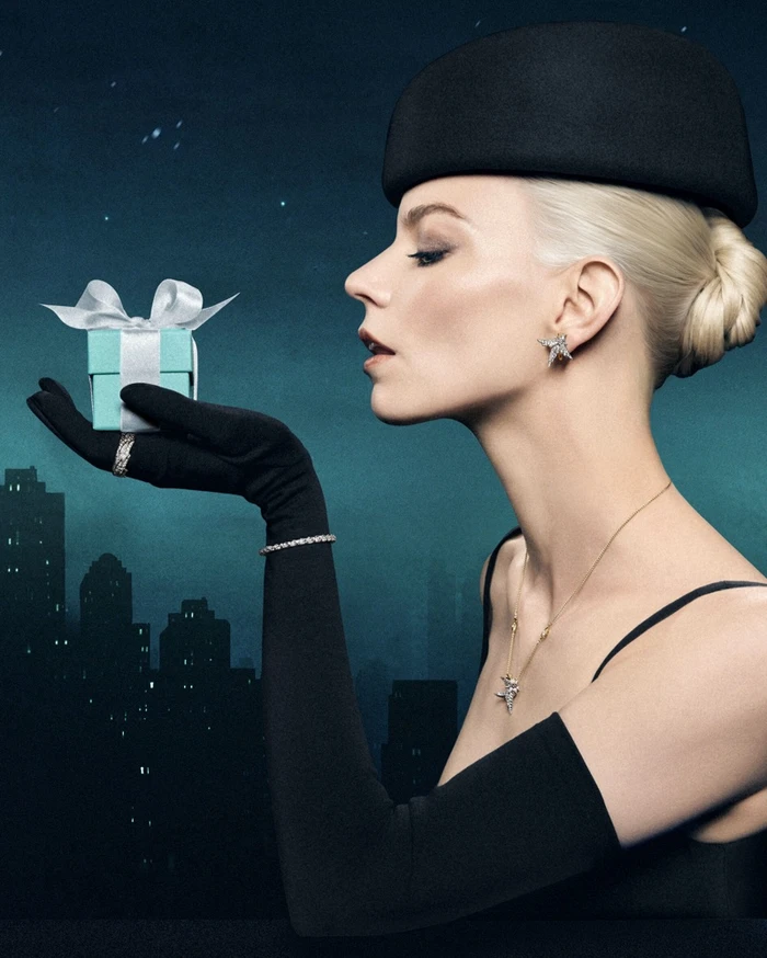 Anya Taylor-Joy wearing a black pillbox hat and opera gloves, holding a small turquoise gift box with a white ribbon.