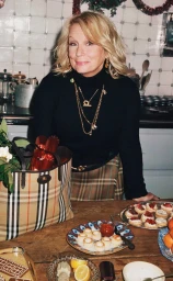 Jennifer Saunders wearing a black turtleneck and plaid skirt standing in a kitchen for Burberry photographed by Angelo Pennetta.