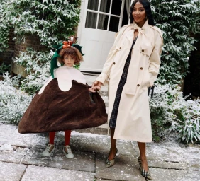 Naomi Campbell in a beige trench coat holding hands with a child in a Christmas pudding costume outside a snow-covered house