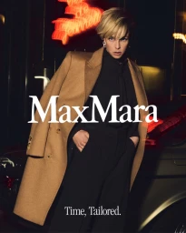 Edie Campbell wearing a camel Max Mara coat over black clothing at night with red neon lights in the background.