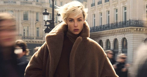 Edie Campbell in a brown teddy coat against a classical street backdrop with motion blurred figures in the foreground