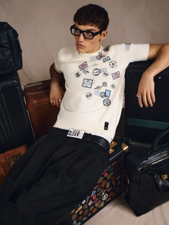 Young adult male model wearing glasses poses surrounded by suitcases and travel bags. The subject wears a white t-shirt and black pants.