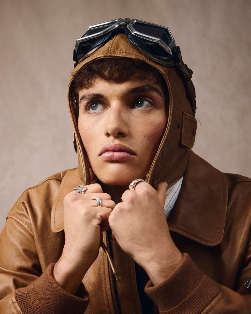 Man in aviator cap and leather jacket gazes upward. Silver rings on fingers. Portrait in a photo studio with neutral background.