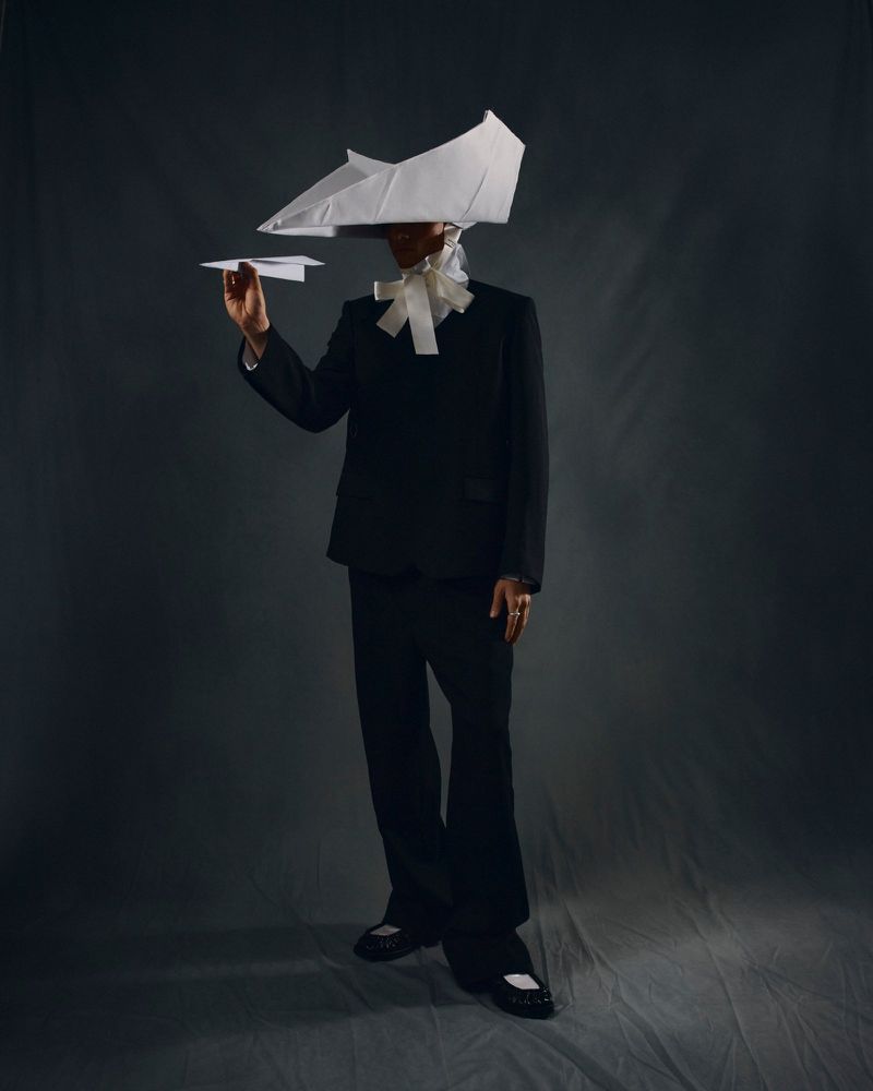 Person in a black suit with a paper plane and paper hat posing in front of a gray backdrop in a studio with creative styling.