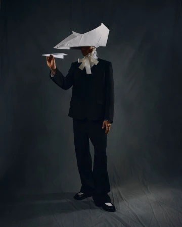 Person in a black suit with a paper plane and paper hat posing in front of a gray backdrop in a studio with creative styling.