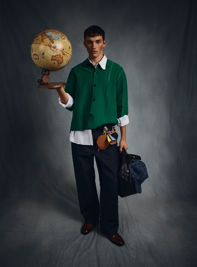A young adult male model poses in a studio with a globe, wearing a green collared shirt, dark trousers, and brown leather shoes, fashion shoot.