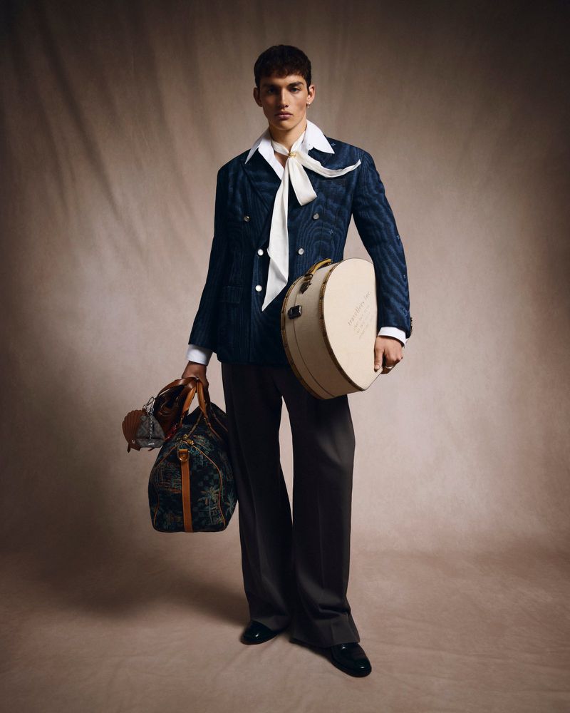 Male fashion model in a patterned blue blazer, grey pants, and white neck scarf, holding a round hat box and duffel bag.