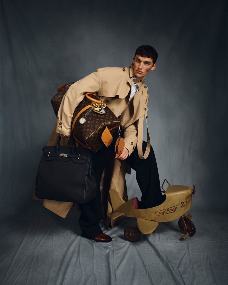 Male fashion model poses in a tan trench coat, carrying a black bag and a brown Louis Vuitton bag with a child's airplane toy.
