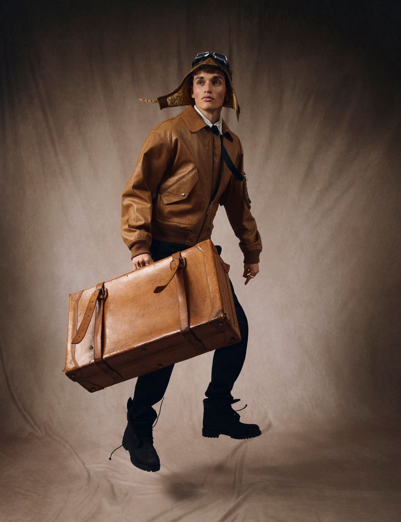 Male model in vintage aviator attire and flight cap leaps with a tan leather suitcase against a neutral studio backdrop, fashion shoot.