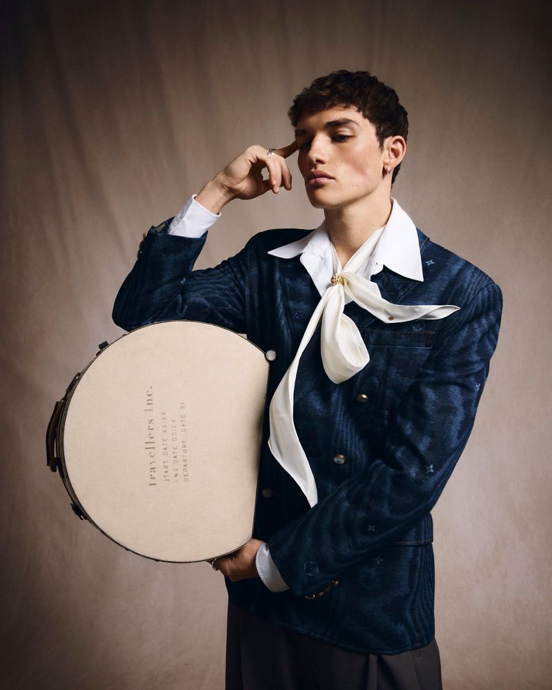 Young adult male poses in a blue blazer, white collared shirt, and cream-colored ascot, holding a drum against a neutral backdrop.
