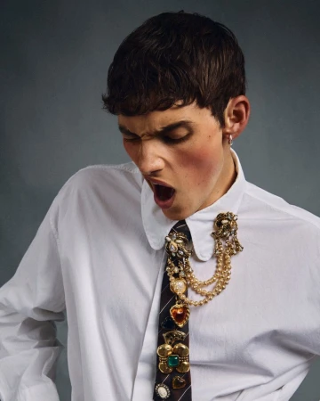 Young adult male in a white dress shirt with a decorated tie, open mouth, looking down against a gray background in a studio shot.
