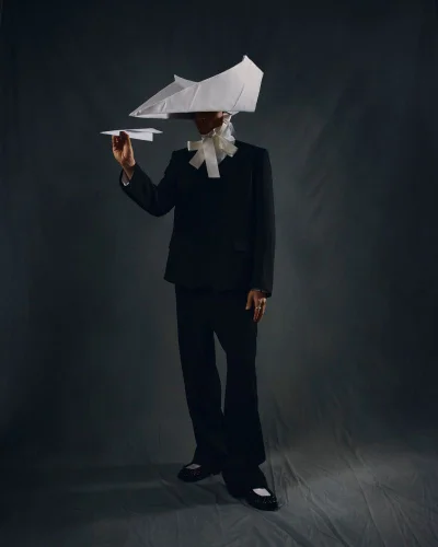 Model in a black suit wearing a large folded white paper hat and holding a paper airplane against a dark textured backdrop.