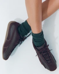 Lila Moss wearing Puma x Madhappy brown suede sneakers and dark green socks against a white seamless background.