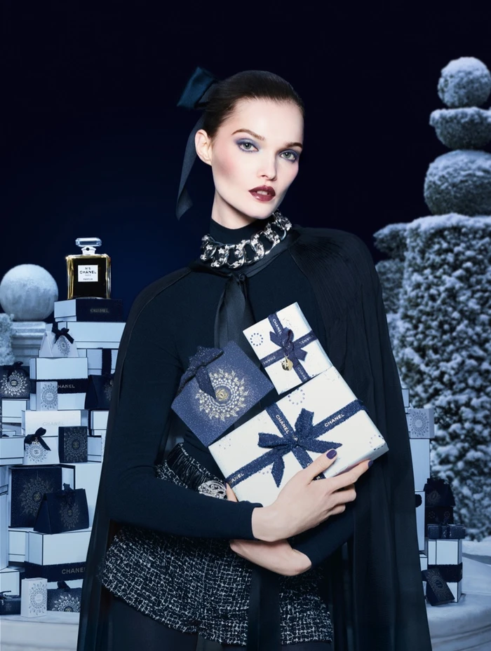 Model Lulu Tenney holding wrapped Chanel holiday gift boxes against a snowy architectural backdrop with perfume bottles.