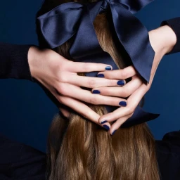 Model with long brown hair tied with a large dark satin bow, hands clasped behind head showing blue nail polish against a solid blue background