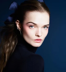Model Lulu Tenney in a dark turtleneck wearing a blue hair bow and sparkling ear cuff against a navy background for Chanel Holiday 2025.