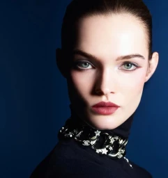 Close-up portrait of model Lulu Tenney wearing a dark turtleneck and statement choker necklace against a deep blue background.