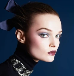 Model Lulu Tenney in Chanel Holiday 2025 campaign with blue eyeshadow, dark lips, hair bows, and a chunky silver chain necklace against a dark blue background.
