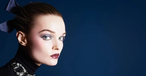 Model Lulu Tenney with blue eyeshadow and a dark bow in her hair against a solid blue studio backdrop for Chanel Holiday 2025.
